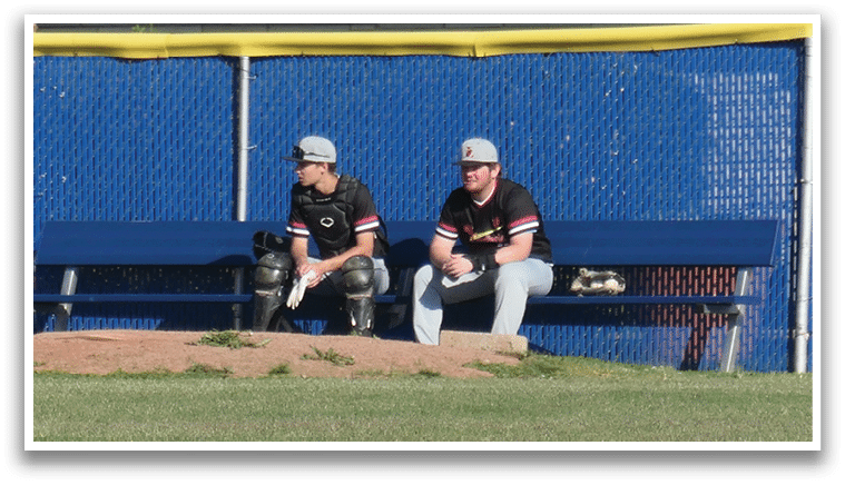 Two men sitting on a bench in front of a baseball field. Description generated by AI
