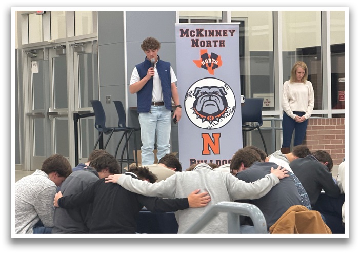A group of people are gathered in a room, with some sitting on chairs and others standing. A man is speaking into a microphone, and the group appears to be listening attentively. The scene takes place in a school gymnasium, with a banner visible in the background. Contenido generado con IA