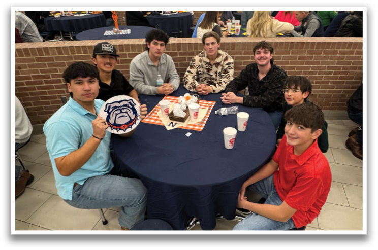 A group of young men and women are sitting around a table, posing for a picture. They are wearing red and orange shirts. Contenido generado con IA
