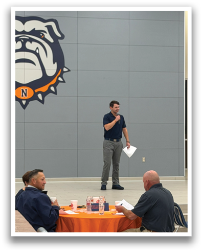 A man is standing in front of a group of people, giving a presentation. The group is seated around a table, with a few cups and bottles placed on it. The man appears to be holding a piece of paper, possibly a speech or presentation notes. Contenido generado con IA