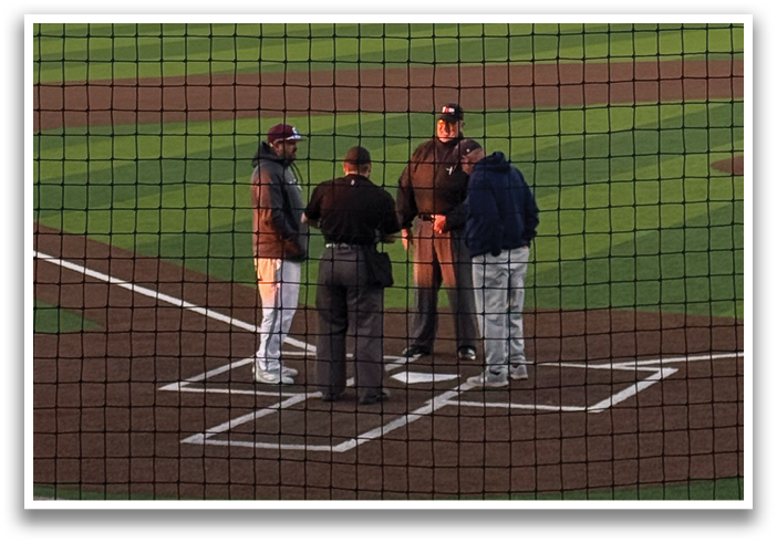 A group of baseball players wearing blue and grey uniforms are standing on a field. They are engaged in conversation with a man in a blue shirt, who is likely a coach or an official. The players are equipped with baseball gloves, and a baseball bat is visible in the scene. Contenido generado con IA