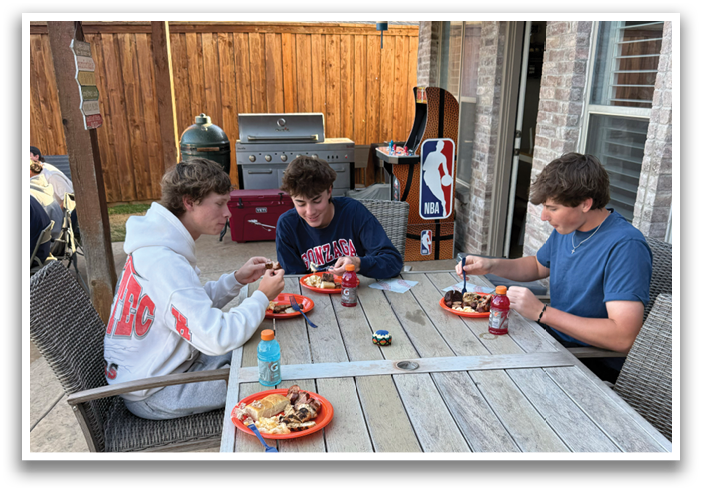 Three people are sitting around a wooden dining table, enjoying a meal together. They have plates of food and drinks in front of them, including cups and bottles. The table is surrounded by chairs, and there is a couch nearby. The scene appears to be an outdoor gathering, with a patio table and chairs set up for the guests. Contenido generado con IA