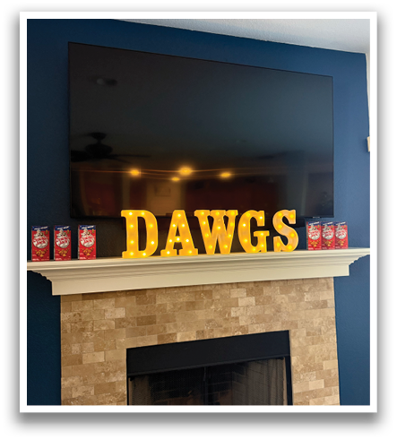 A flat screen TV is mounted on a wall above a fireplace. The fireplace has a brick design and is adorned with a sign that reads “Dogs." There are several cans of soda placed around the fireplace, adding to the cozy atmosphere. Contenido generado con IA