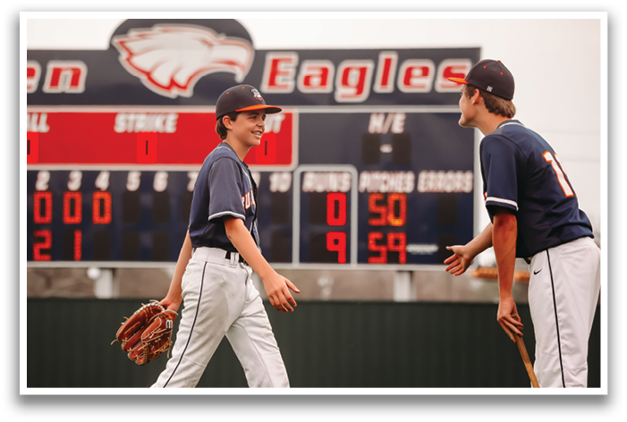 Two baseball players are standing on a field, one holding a baseball glove. They are positioned in front of a scoreboard displaying the current score. Contenido generado con IA