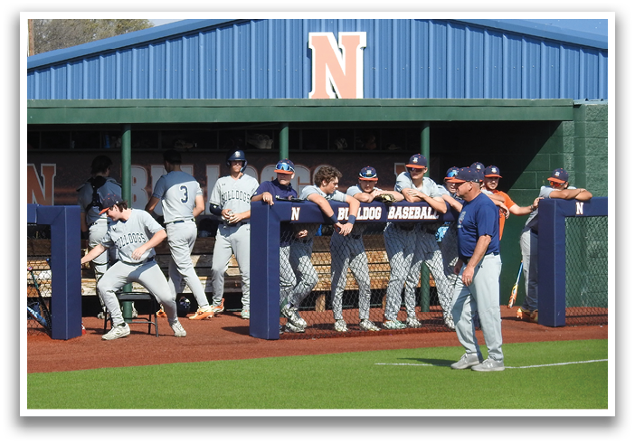 Baseball players in uniforms are gathered on the field, some standing and others sitting on benches. A man is standing near the players, possibly a coach or teammate. The scene takes place in front of a blue and white building, likely a dugout. Contenido generado con IA