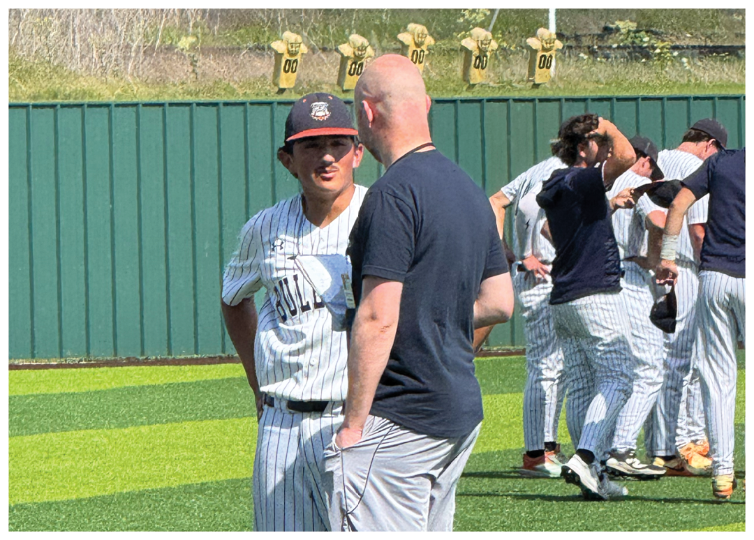 Two men are standing on a baseball field, engaged in conversation. One man is wearing a baseball uniform, while the other is not. Contenido generado con IA