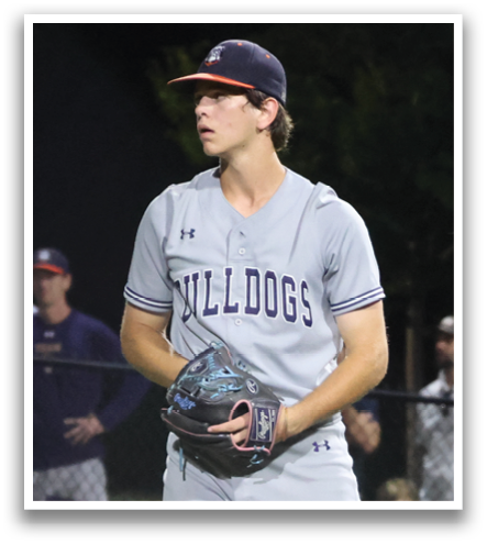 A baseball player is standing on the pitcher's mound, wearing a grey uniform and a blue hat. He is holding a baseball in his hand, preparing to throw it. There are several other people in the scene, some of whom are standing near the fence, watching the game. Contenido generado con IA