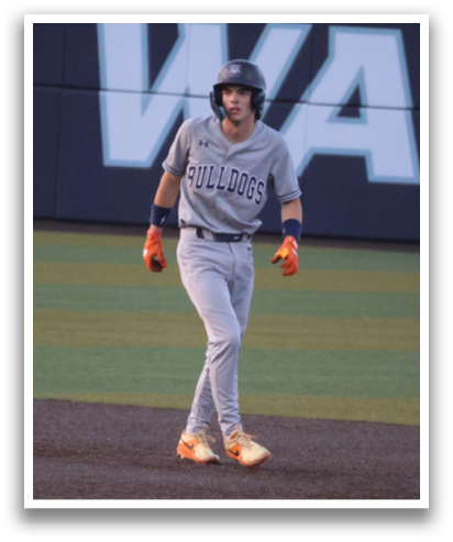A baseball player is standing on the field, wearing a grey uniform and holding a bat. The player is positioned near a base, ready to run. There are other people in the background, possibly teammates or opponents. Contenido generado con IA