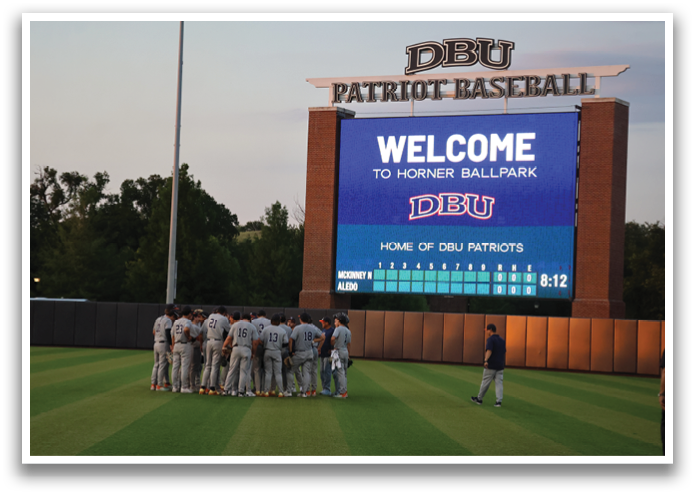 A group of baseball players huddled together on a field in front of a large scoreboard. Contenido generado con IA