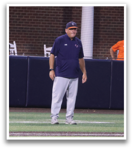 A baseball coach stands on the field, wearing a blue shirt and a hat. Contenido generado con IA