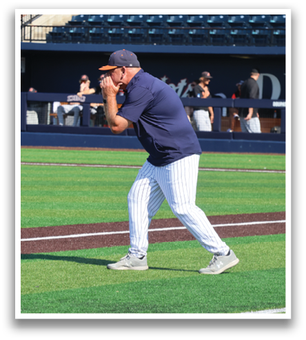 A baseball coach is standing on the field, wearing a blue shirt and a cap. He is talking on a cell phone. Contenido generado con IA