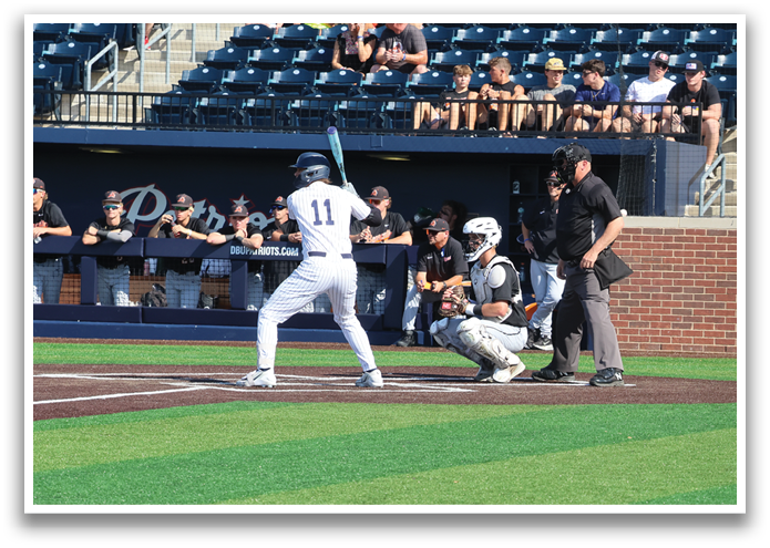 A baseball player is at bat, holding a bat and getting ready to swing. The catcher and umpire are positioned behind him, and there are several other players on the field. The scene takes place in a stadium with a crowd of spectators watching the game. Contenido generado con IA