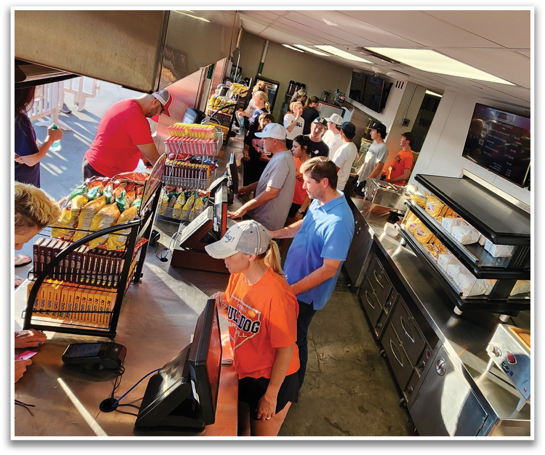 A group of people are standing in a kitchen, with some of them holding beers. There are several bottles and cups on the counter, and a refrigerator is visible in the background. Contenido generado con IA
