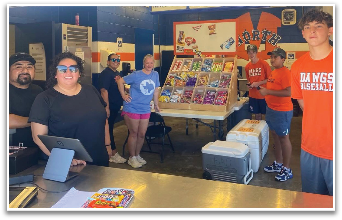 A group of people are standing in a room with a table full of food. A man in an orange shirt is standing next to a man in a black shirt. There are several chairs in the room, and a refrigerator is visible in the background. Contenido generado con IA