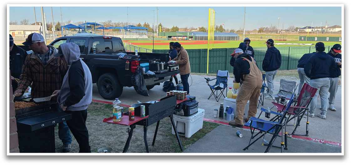 A group of people are gathered around a grill, preparing food. There are several chairs and a dining table set up nearby. A baseball game is being played in the background, with a baseball bat visible. A truck is parked nearby, and a few bottles can be seen on the ground. Contenido generado con IA