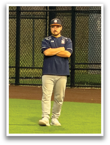 A man wearing a blue shirt and khaki pants stands on a baseball field. Contenido generado con IA