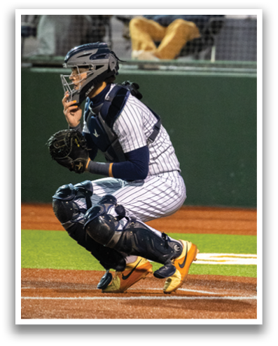 A baseball catcher is kneeling on the field, wearing a baseball glove. The catcher is positioned behind the home plate, ready to catch the ball. A baseball umpire is standing nearby, observing the game. Contenido generado con IA