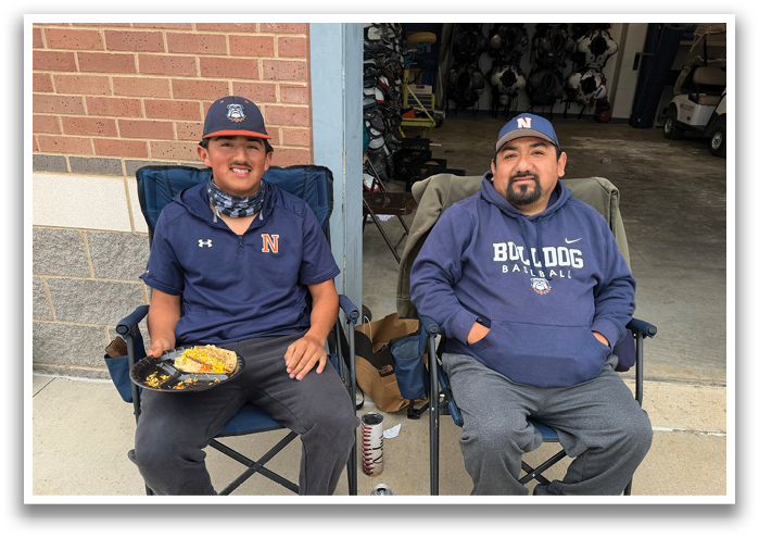 Two men are sitting in chairs outside, eating food. One man is wearing a blue shirt with the word “North" on it. Contenido generado con IA
