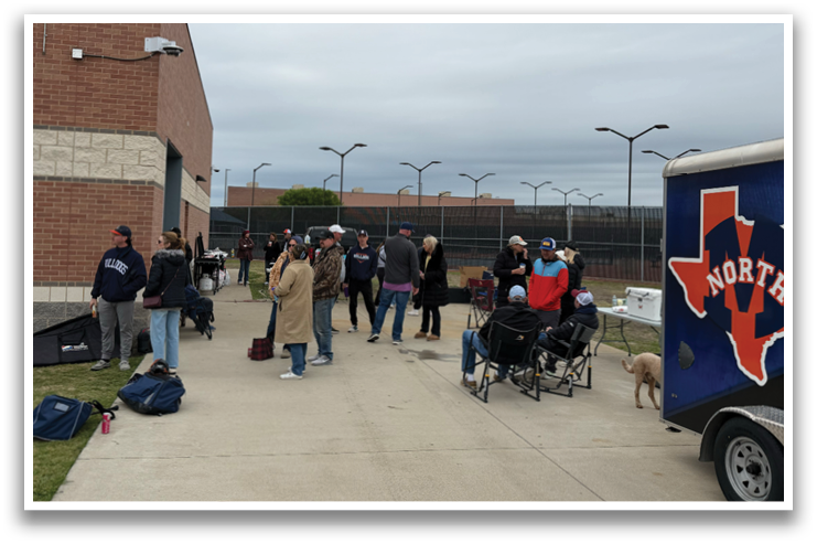 A group of people are standing outside a building, some of them are holding suitcases. A dog is also present in the scene. A blue truck is parked nearby. Contenido generado con IA