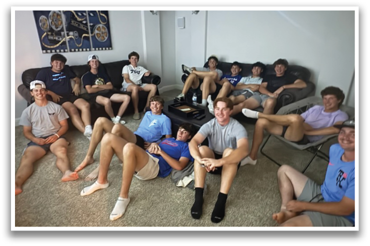 A group of young men are sitting in a living room, some on a couch and others on chairs. They appear to be relaxed and enjoying each other's company. Contenido generado con IA