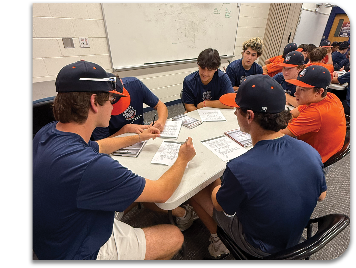 A group of men are sitting around a table, wearing baseball caps and shirts. They are engaged in a discussion or possibly a game. There are several chairs around the table, and a whiteboard is visible in the background. Contenido generado con IA