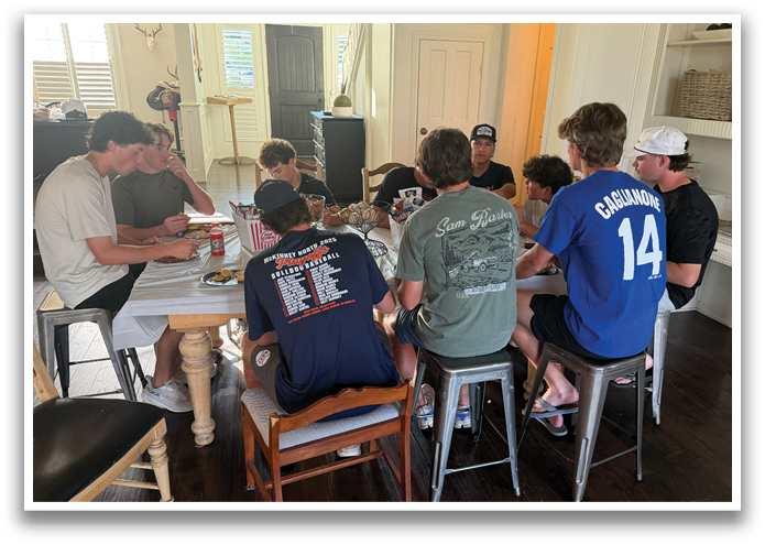 A group of people are sitting around a table, eating and drinking. They are wearing blue shirts and are engaged in conversation. There are several chairs around the table, and a potted plant is visible in the background. Contenido generado con IA