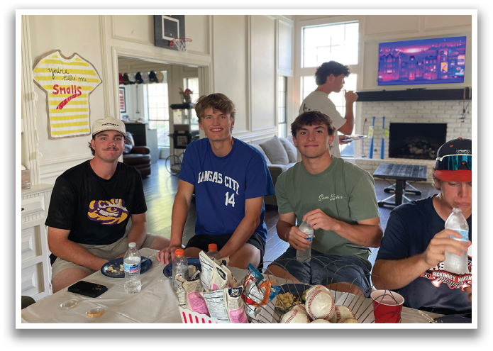 Four young men are sitting around a table, enjoying a meal together. They are holding drinks and snacks, and there are sports balls on the table. A TV is mounted on the wall above them. Contenido generado con IA