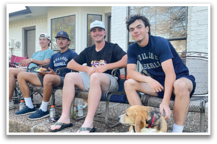 Four young men are sitting on a bench, with a dog sitting between them. They are all wearing baseball caps. Contenido generado con IA