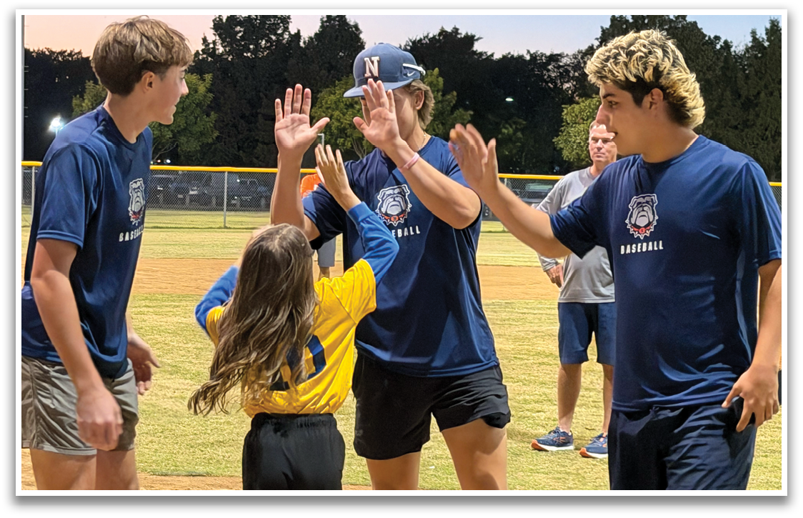 Three young men and a young girl are on a baseball field. The girl is holding up her hand to the two men, who are also holding up their hands. Contenido generado con IA