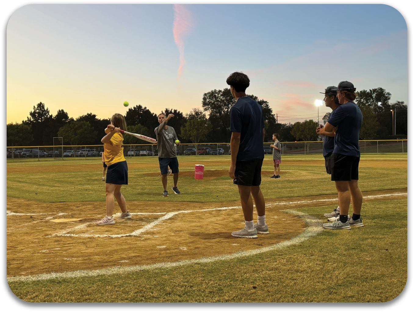 A group of people are playing baseball on a field. A woman is throwing a ball towards a man who is holding a bat, ready to hit the ball. Other people are watching the game from the sidelines. Contenido generado con IA