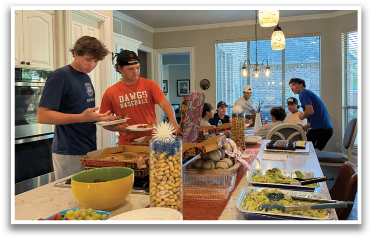 A group of people are gathered in a large kitchen, eating and socializing. The table is filled with plates of food, including salad and pizza. There are several chairs around the table, and a bowl of fruit is also present. The kitchen is well-equipped with an oven, sink, and refrigerator. The atmosphere appears to be casual and friendly, with everyone enjoying their time together. Contenido generado con IA