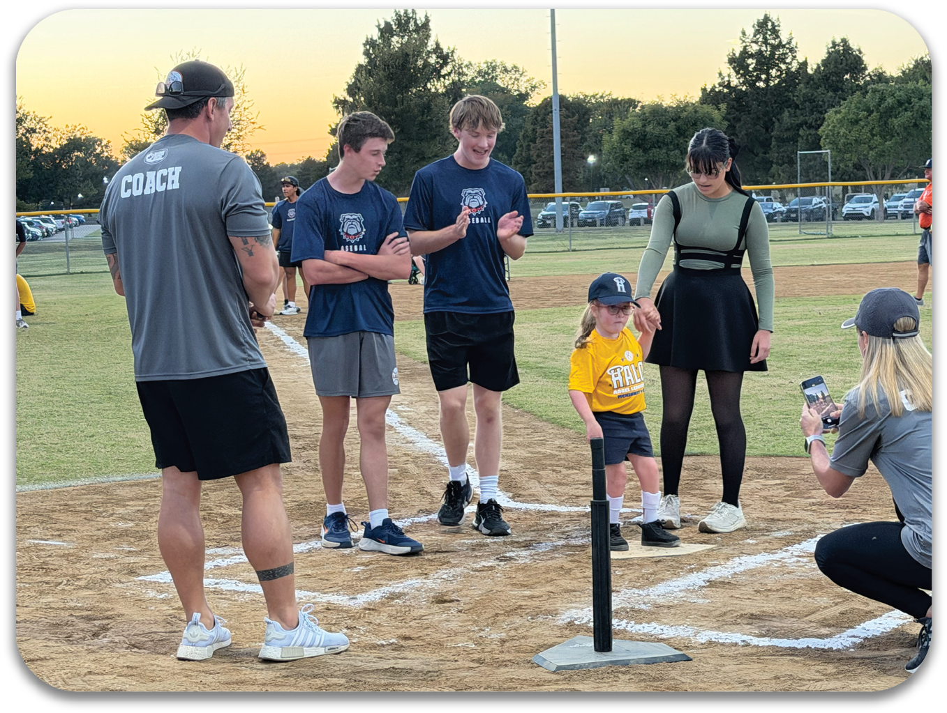 A group of people, including a woman and a child, are standing on a baseball field. They are posing for a picture in front of a base. There are several cars parked in the background. Contenido generado con IA