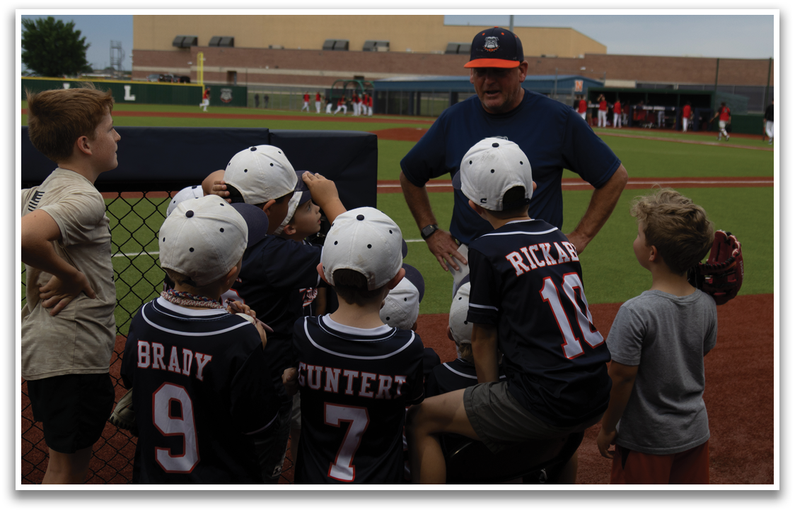 A group of young boys wearing baseball uniforms are gathered around a man, possibly a coach, who is giving them instructions. Contenido generado con IA