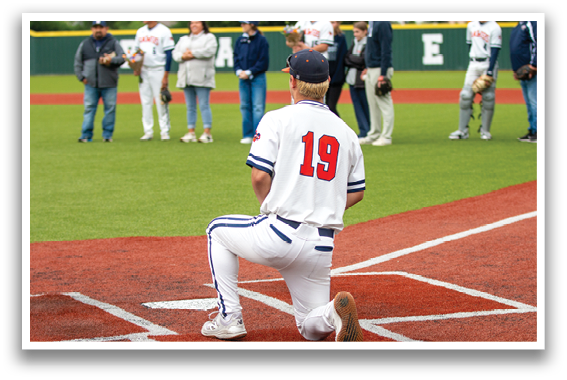 A baseball player is kneeling on the pitcher's mound, wearing a white uniform and a blue hat. Contenido generado con IA
