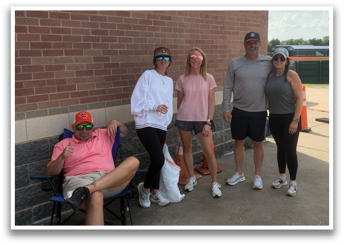 A group of people are standing outside a building, posing for a picture. There are five people in total, with two women and three men. They are all wearing sunglasses, and one of the women is wearing a pink shirt. The group is standing in front of a brick wall, and there is a chair nearby. Contenido generado con IA