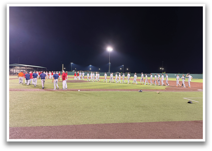 A group of baseball players are standing on a field, some of them wearing red and white uniforms. They are lined up, possibly waiting for their turn to bat or for the game to start. The players are standing in a line, with some of them closer to the foreground and others further back. The scene is set at night, with the players and the field illuminated by artificial light. Contenido generado con IA