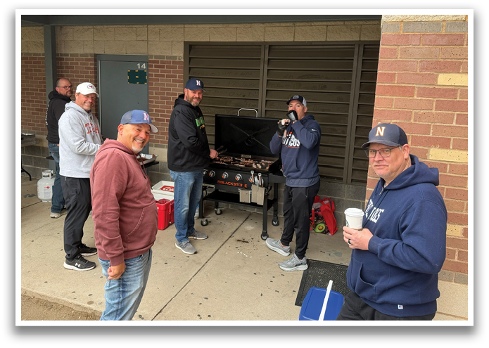 A group of men are standing around a grill, smiling and enjoying the outdoor cooking event. Contenido generado con IA