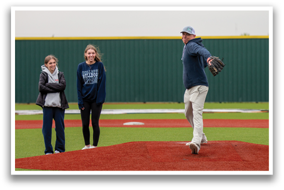 A man in a blue shirt and white pants is pitching a baseball on a field. Two women are watching from the sidelines. Contenido generado con IA