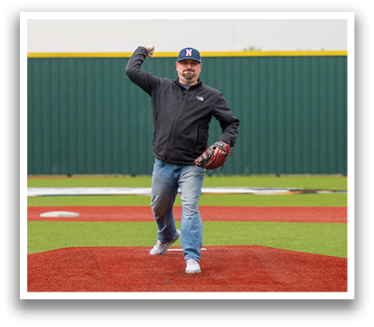 A man in a black jacket and blue hat is throwing a baseball on a baseball field. A woman is standing behind him, clapping. Contenido generado con IA