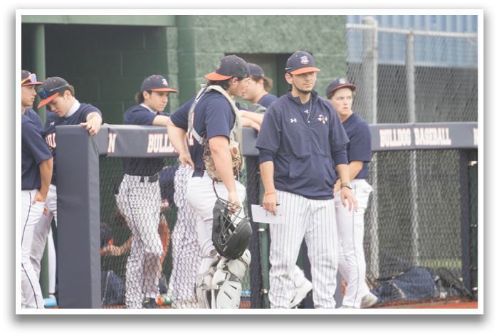 Baseball players are standing on the field, some of them wearing blue and orange uniforms. They are gathered near the dugout, possibly waiting for their turn to play or discussing the game. Contenido generado con IA