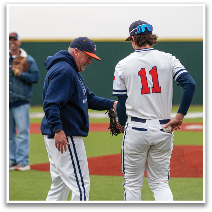 Two men are standing on a baseball field, one of them wearing a baseball uniform. The man in the uniform is wearing a blue and white hat and a blue and white jersey. The other man is wearing a blue jacket and a blue hat. They are shaking hands, possibly after a game. Contenido generado con IA