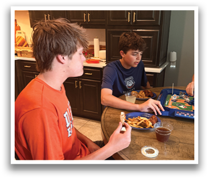 Two boys are sitting at a table with plates of food. They are playing a game on a tablet. Contenido generado con IA