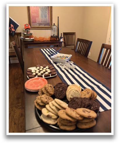 A table is set with a variety of desserts, including cookies, cakes, and candies. The table is adorned with a blue and white striped tablecloth and a bowl of candy. There are several chairs surrounding the table, and a flag can be seen in the background. Contenido generado con IA