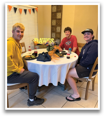 Three people are sitting around a table with food and drinks. They are wearing shorts and appear to be enjoying a meal together. Contenido generado con IA