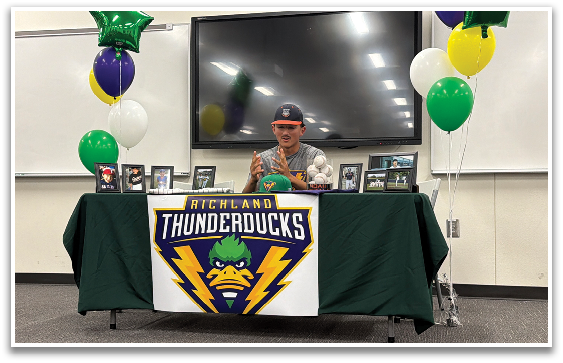 A man stands behind a table with a green and yellow tablecloth, posing for a picture. The table is decorated with a banner and balloons. Contenido generado con IA
