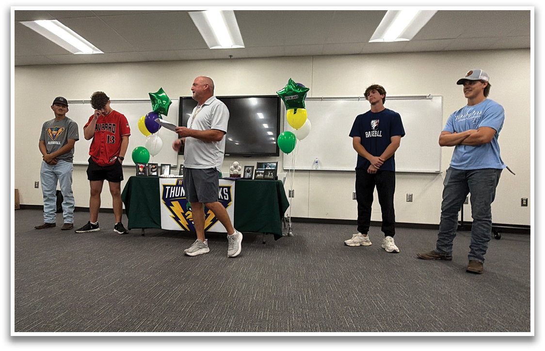 A group of young men stand in a room, some wearing shorts. A man in a white shirt is speaking to the group. There is a table with a laptop and a TV in the background. Contenido generado con IA