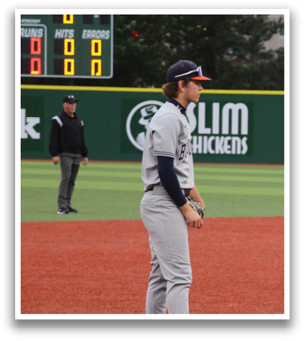 A baseball player stands on the field, wearing a grey uniform and a blue shirt. He is holding a baseball bat and looking at the ground. Contenido generado con IA