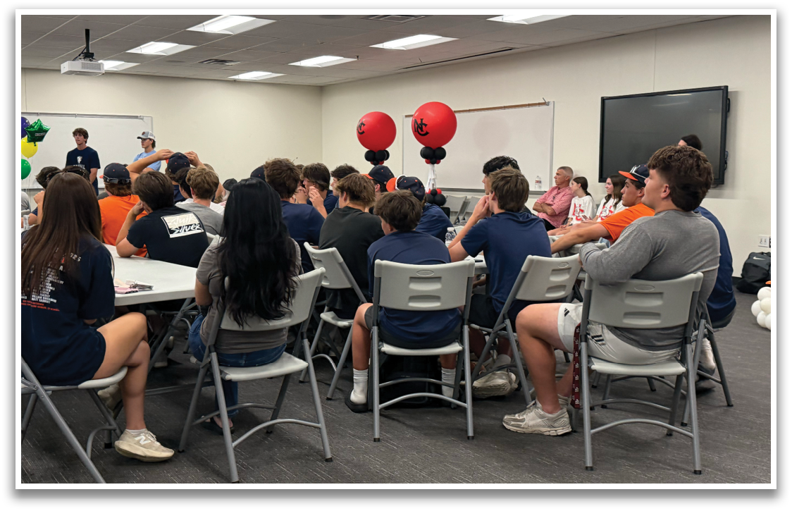 A group of people are sitting in a room, some of them are wearing blue shirts. A boy is sitting in the front row. There are chairs and a dining table in the room. A TV is mounted on the wall. Contenido generado con IA