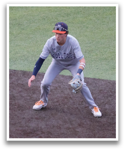 A baseball player is standing on a field, wearing a grey uniform and a blue hat. He is holding a baseball glove, ready to catch the ball. Contenido generado con IA