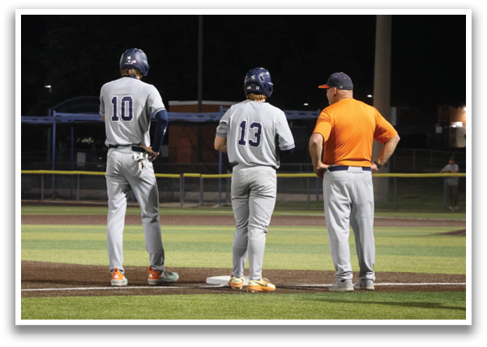 Two baseball players stand on the field, one wearing a blue shirt and the other wearing an orange shirt. A coach is also present, observing the players. Contenido generado con IA